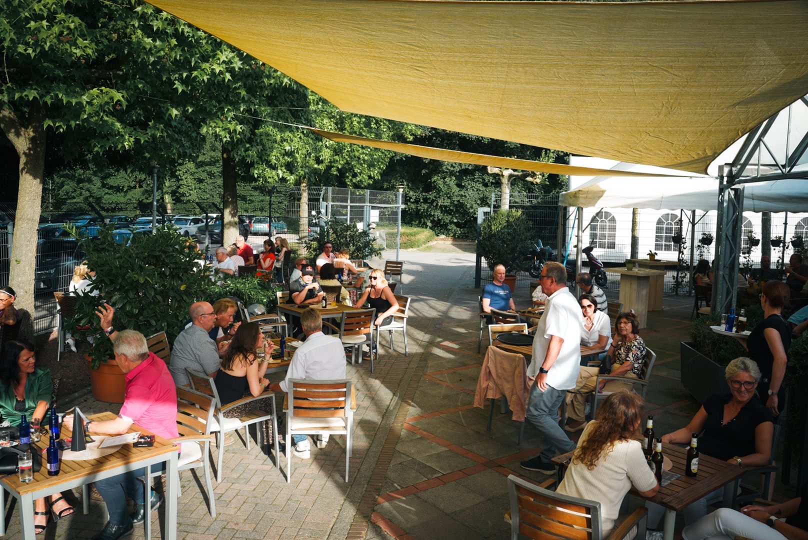 Voller Biergarten: Gäste an Holztischen unter dem Sonnensegel, dahinter die Bäume