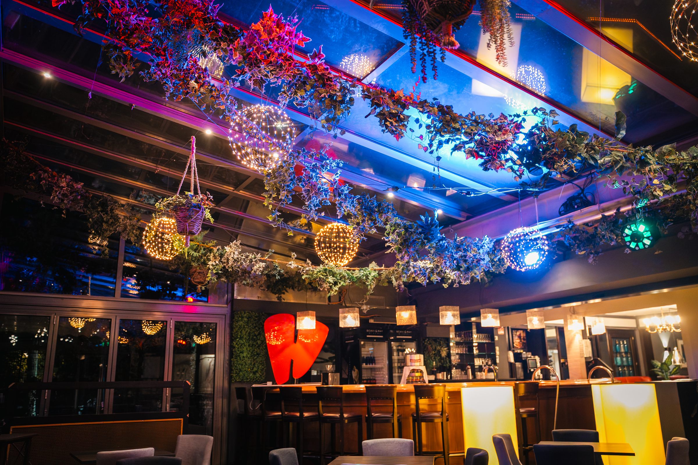 Der Glaspalast bei Nacht: buntes Licht, Lichterkugeln und Blätterdach unter dem Glasdach, dahinter die Bar