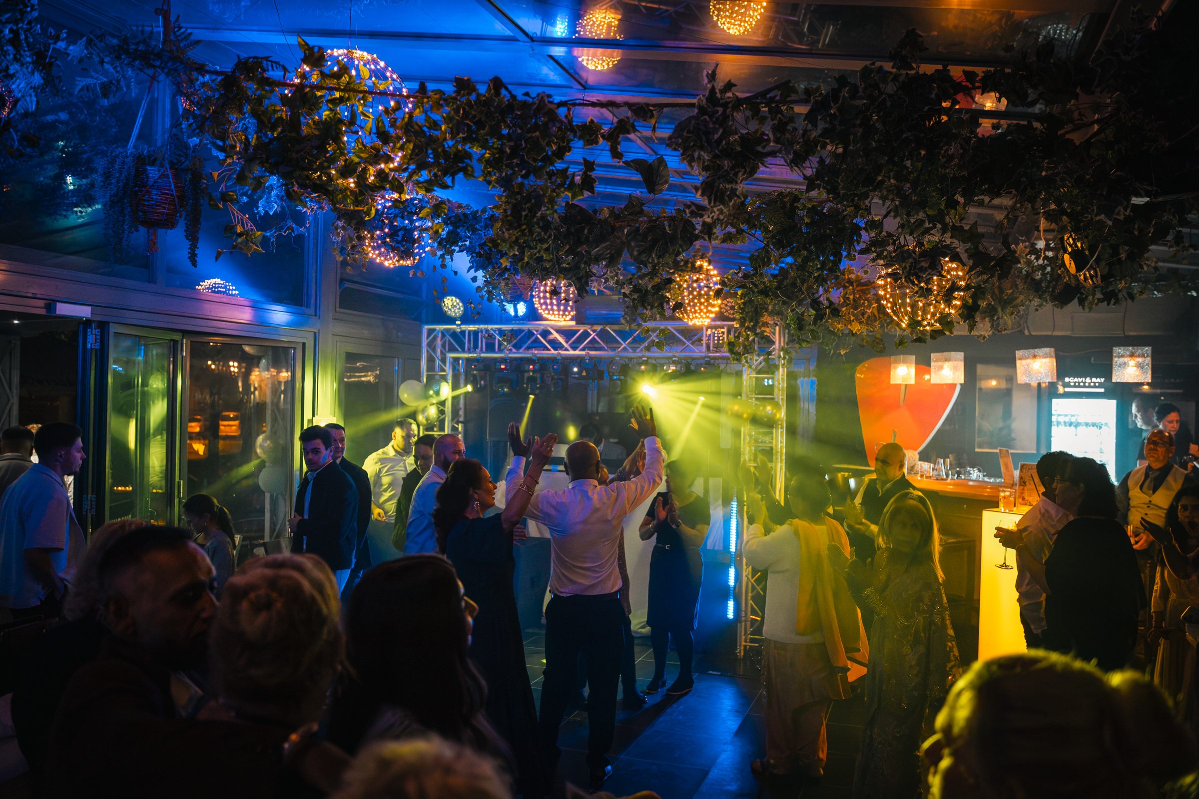 Hochzeitsfeier im Glaspalast: Gäste tanzen unter Lichterkugeln und grünem Blätterdach, dahinter die Lichtshow